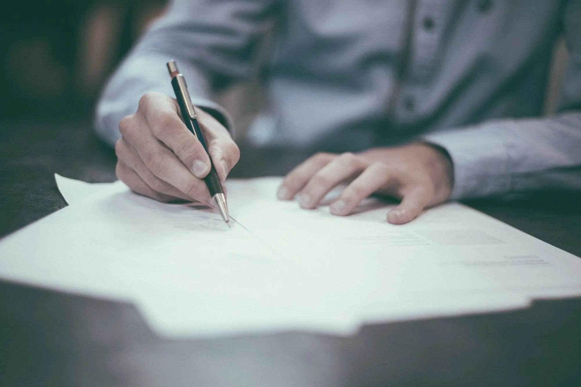 A person in a dress shirt signs a paper form with a pen at a desk.