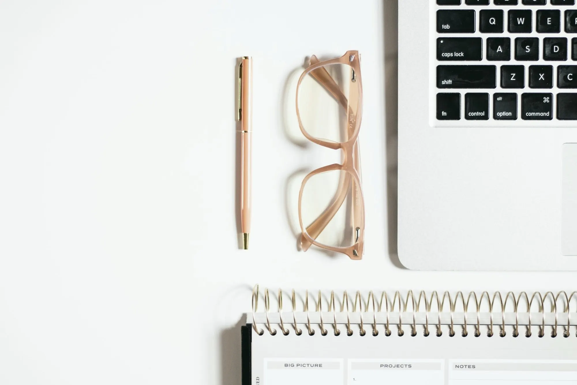 A laptop keyboard, eyeglasses, a pen and a spiral notepad on a white desk.