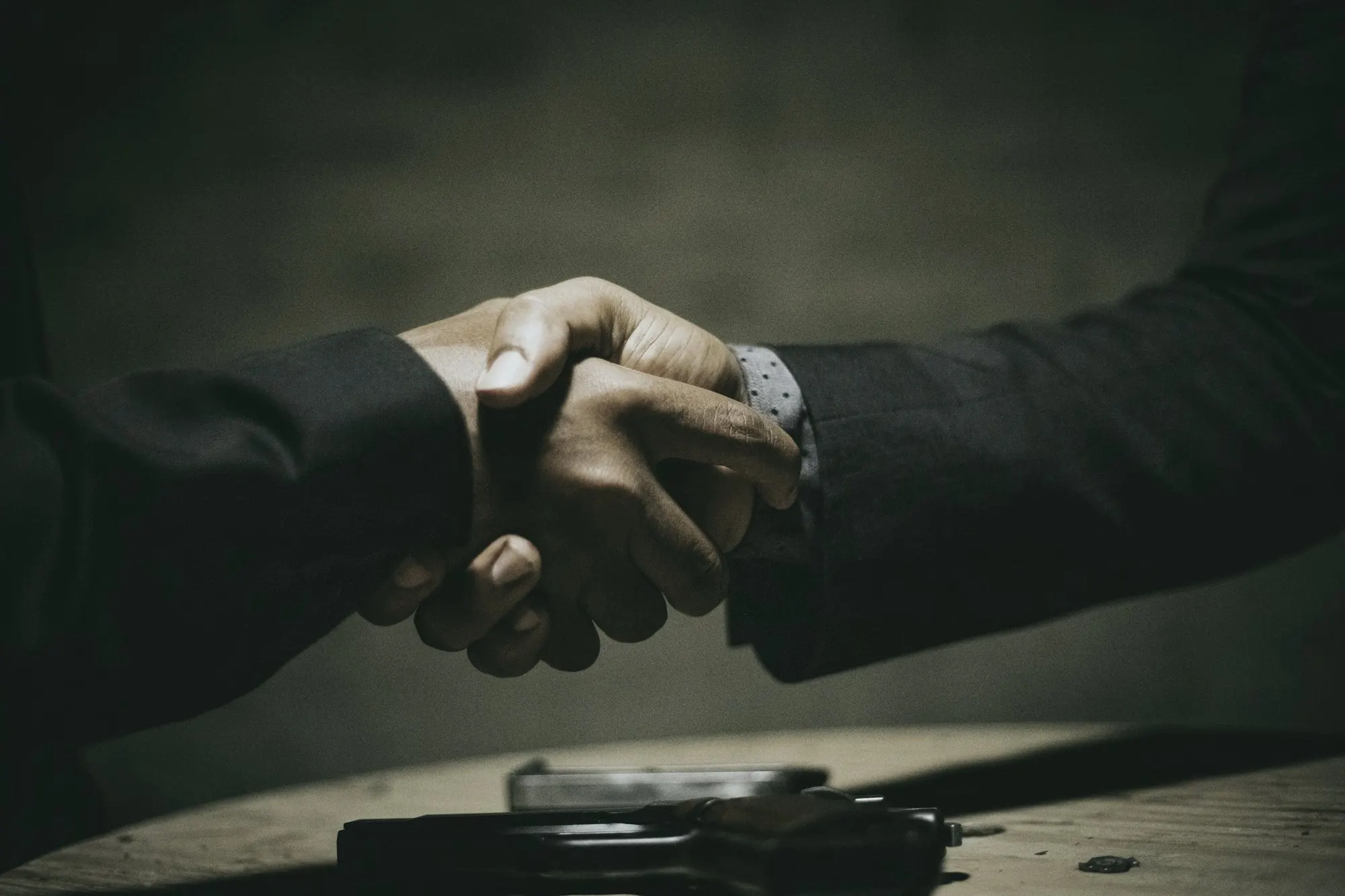 Two people in dark suits shake hands above a wooden table in low light.