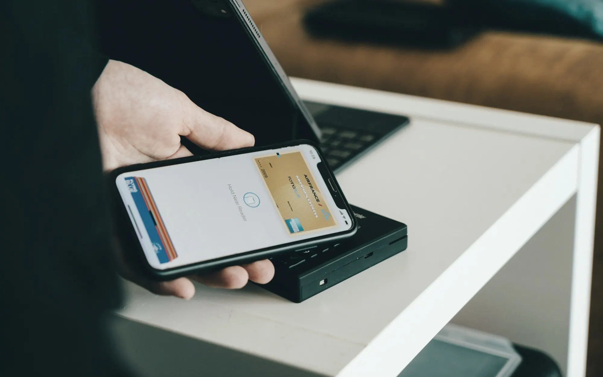 A hand holds a smartphone showing a digital wallet card over a contactless payment reader.