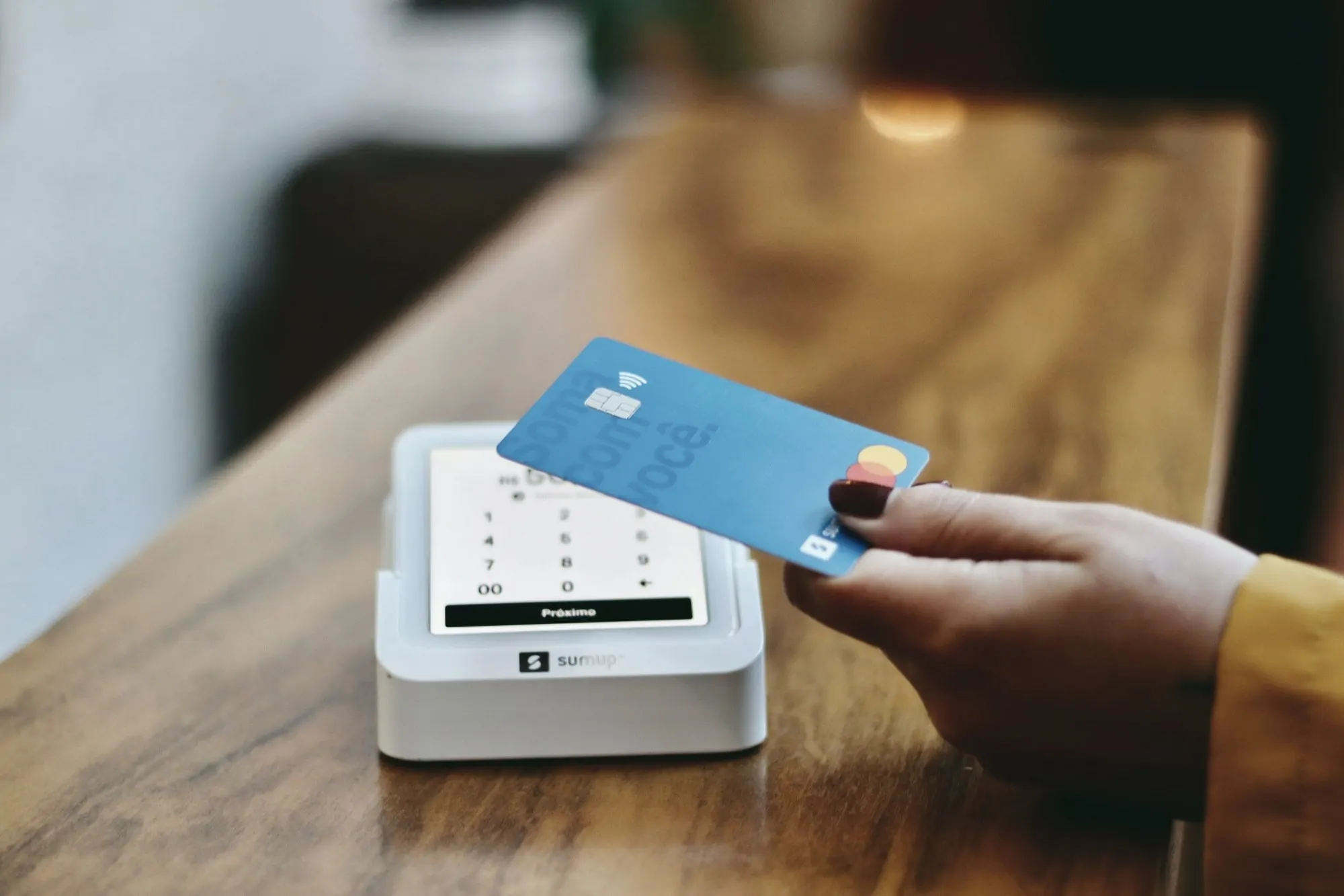 A hand holds a blue credit card over a white countertop card terminal on a wooden counter.