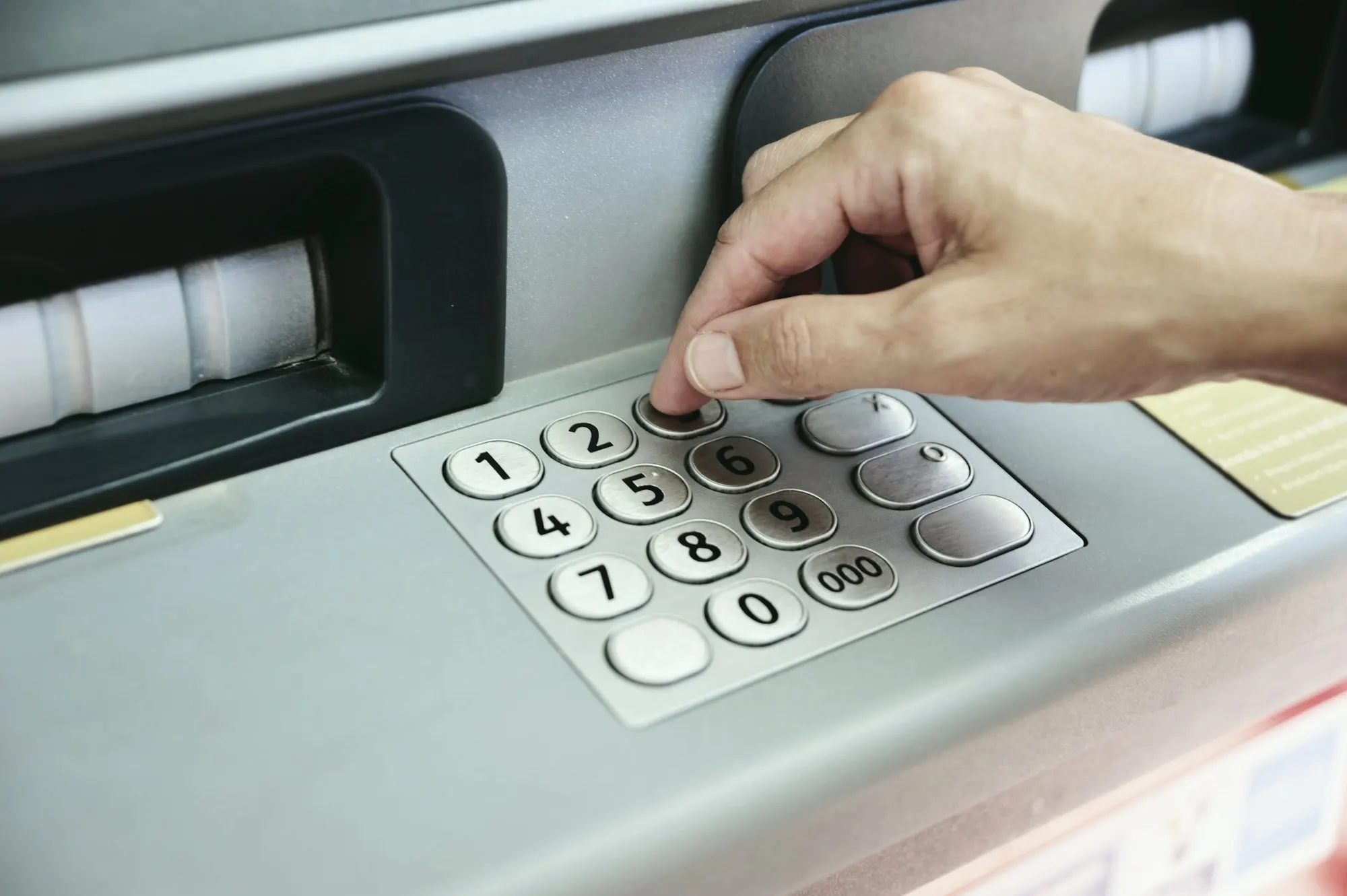 A hand pressing a key on a metal ATM keypad beside the card slot.