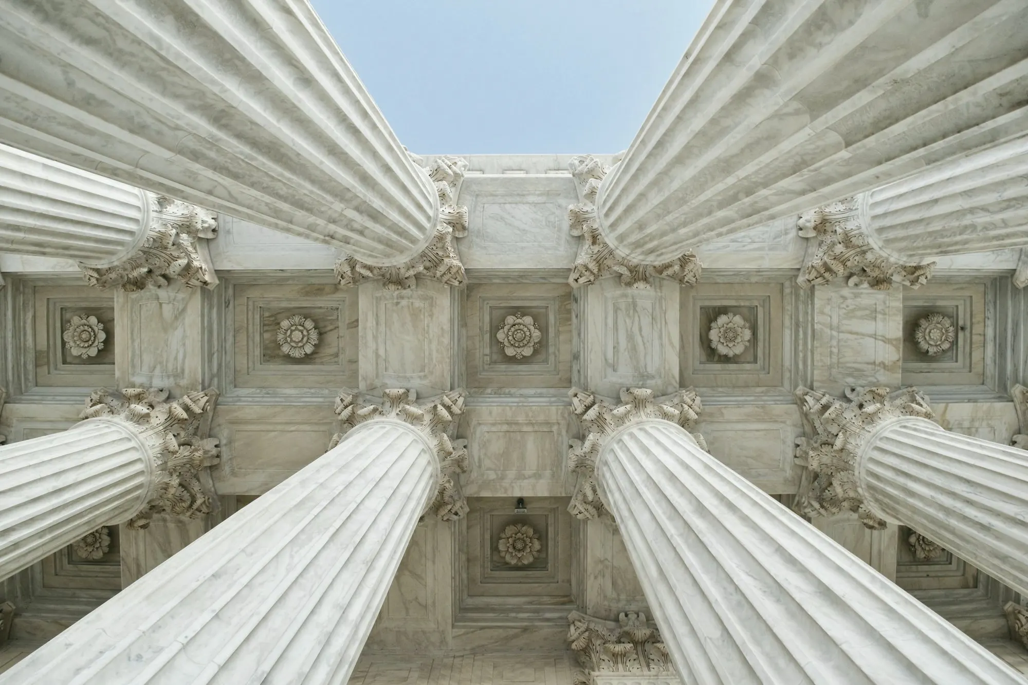 Looking straight up between tall white marble columns toward a coffered stone ceiling and sky.