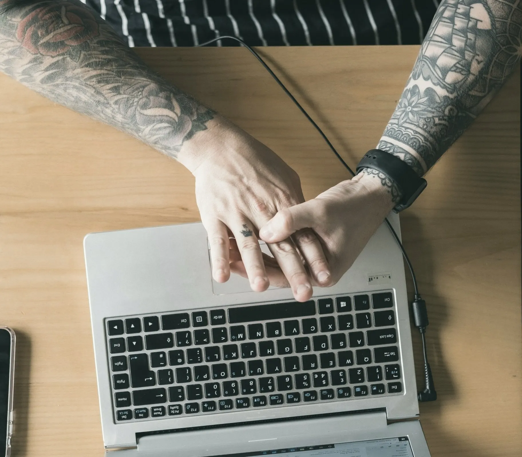 Overhead view of tattooed hands resting on an open laptop on a wooden desk.