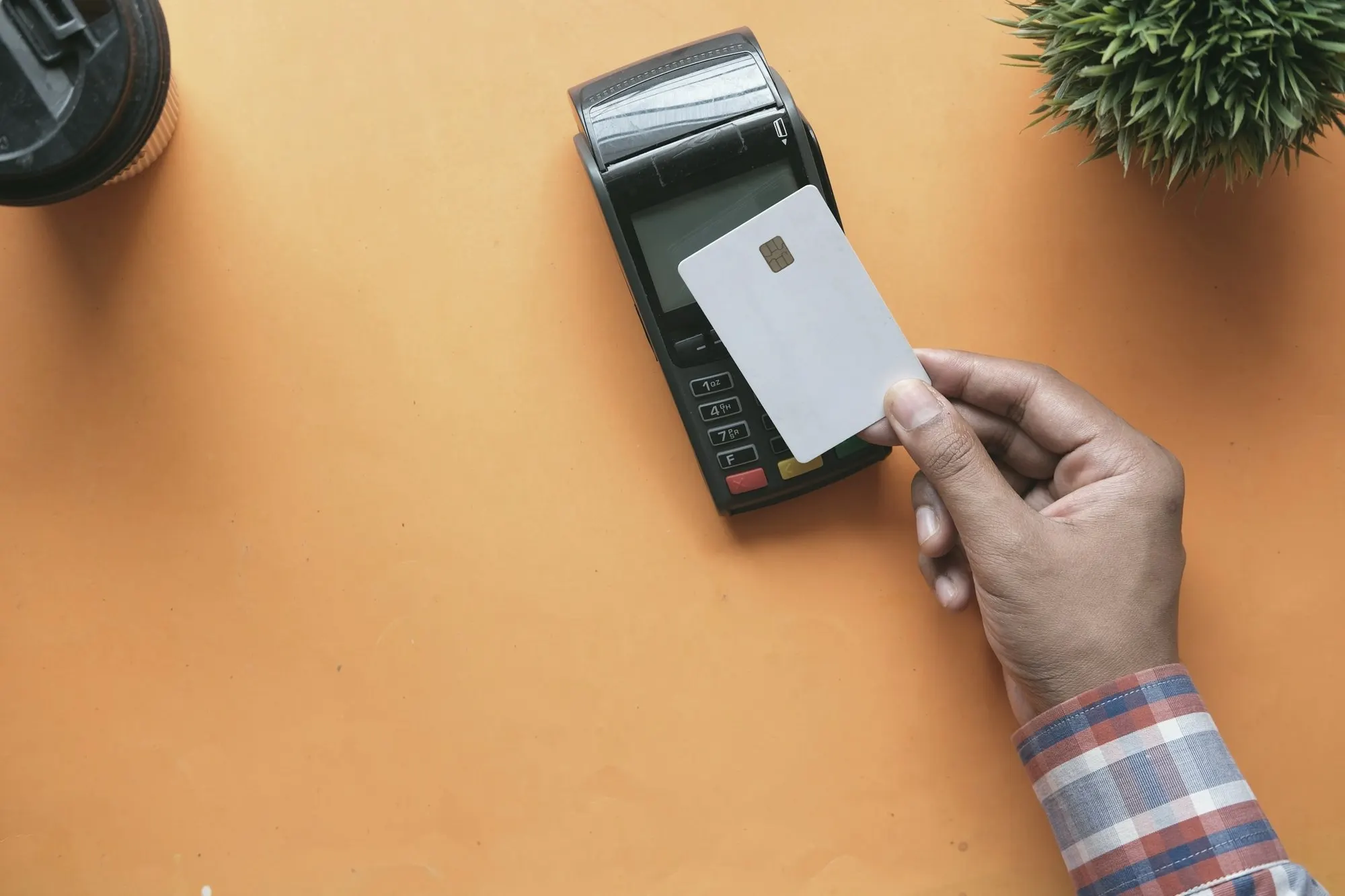A hand holds a blank chip card beside a payment terminal on an orange surface.